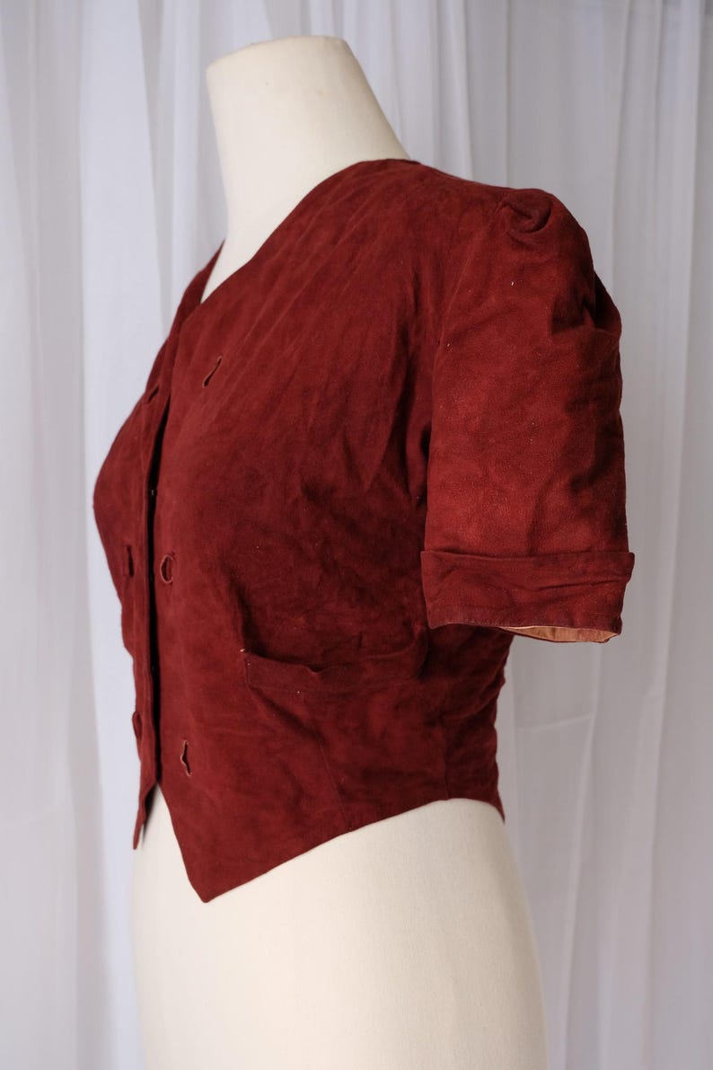 K&ouml;nnte beinhalten: Burgunderfarbene Wildlederjacke mit kurzen &Auml;rmeln und Knopfleiste. Die Jacke hat einen V-Ausschnitt und eine kleine Tasche auf der linken Seite. Die &Auml;rmel sind umgeschlagen und die Jacke wird auf einer Schaufensterpuppe pr&auml;sentiert.