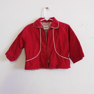 May include: A vintage red jacket with white trim and a zipper closure. The jacket has a collar and long sleeves. The interior lining has a patterned design. The jacket is hanging on a white hanger against a white wall.