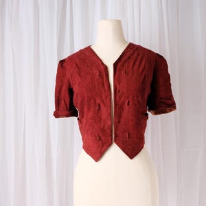 K&ouml;nnte beinhalten: Kurz&auml;rmelige, weinrote Jacke mit V-Ausschnitt und dekorativen N&auml;hten. Die Jacke wird auf einer Schaufensterpuppe vor wei&szlig;em Hintergrund pr&auml;sentiert. Die &Auml;rmel sind umgeschlagen und die Jacke hat eine taillierte Passform.