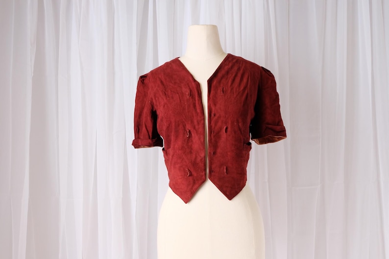 K&ouml;nnte beinhalten: Kurz&auml;rmelige, weinrote Jacke mit V-Ausschnitt und dekorativen N&auml;hten. Die Jacke wird auf einer Schaufensterpuppe vor wei&szlig;em Hintergrund pr&auml;sentiert. Die &Auml;rmel sind umgeschlagen und die Jacke hat eine taillierte Passform.