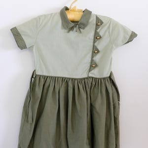 May include: A vintage-style dress in sage green and olive green, hanging on a wooden hanger. The dress features a collared neckline with a small bow, short sleeves, and decorative gold buttons. The skirt is pleated.