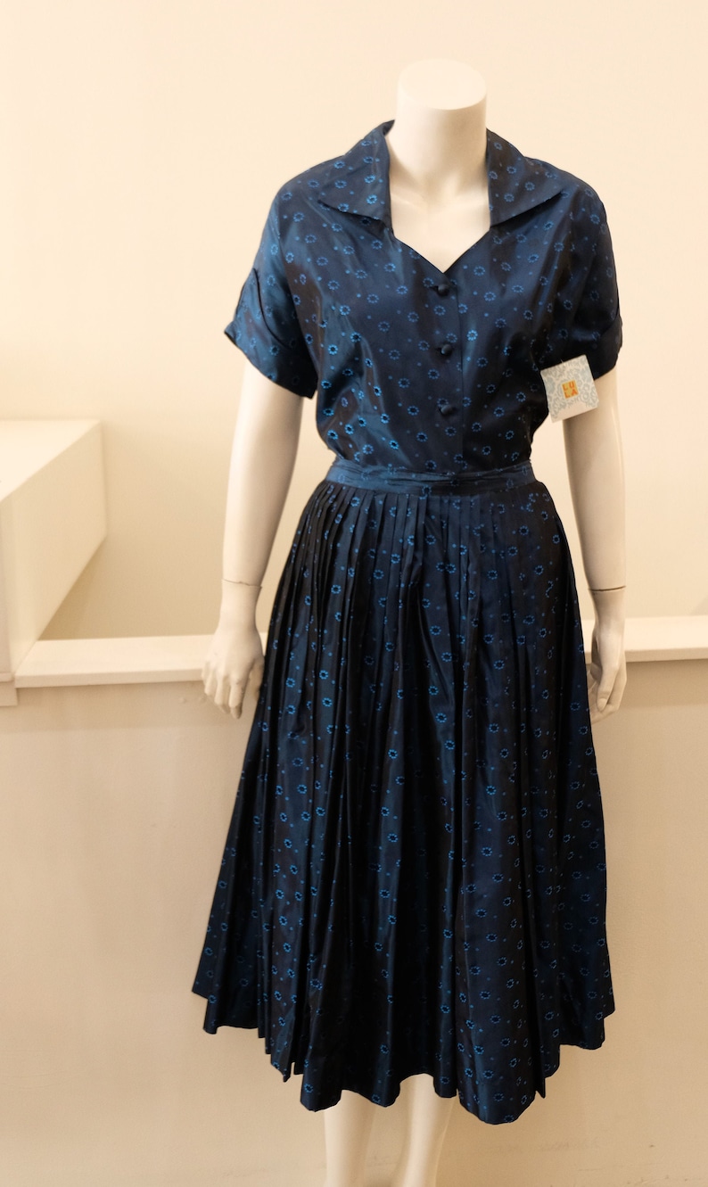K&ouml;nnte beinhalten: Ein marineblaues Vintage-Kleid mit passendem Oberteil und plissiertem Rock. Der Stoff weist ein sich wiederholendes Muster aus kleinen blauen Blumen auf. Das Kleid wird auf einer Schaufensterpuppe pr&auml;sentiert, die seine klassische Silhouette und sein Design zeigt.
