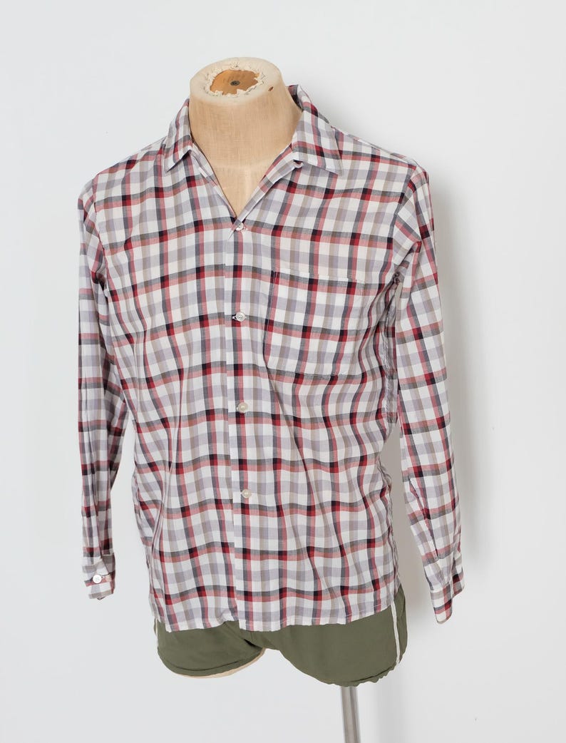 May include: A long-sleeved, button-down shirt with a plaid pattern in red, black, and white. The shirt is displayed on a mannequin and paired with olive green shorts. The shirt has a classic collar and buttoned cuffs.