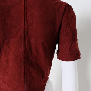 K&ouml;nnte beinhalten: Burgunderfarbene Wildlederjacke mit kurzen &Auml;rmeln und umgeschlagenen Manschetten. Die R&uuml;ckseite der Jacke ist sichtbar und zeigt eine vertikale Naht in der Mitte. Die Jacke wird auf einer wei&szlig;en Schaufensterpuppe pr&auml;sentiert.