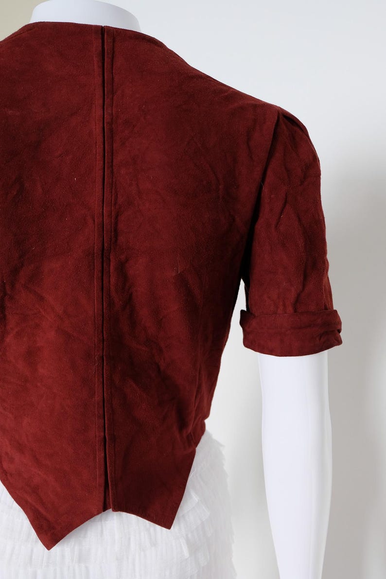 K&ouml;nnte beinhalten: Burgunderfarbene Wildlederjacke mit kurzen &Auml;rmeln und umgeschlagenen Manschetten. Die R&uuml;ckseite der Jacke ist sichtbar und zeigt eine vertikale Naht in der Mitte. Die Jacke wird auf einer wei&szlig;en Schaufensterpuppe pr&auml;sentiert.