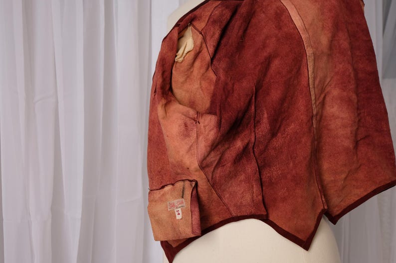 K&ouml;nnte beinhalten: Nahaufnahme einer rotbraunen Lederweste mit einzigartiger, strukturierter Optik. Die Weste hat eine dunkelburgunderfarbene Paspel und eine kleine Tasche mit Etikett. Das Innenfutter ist heller.