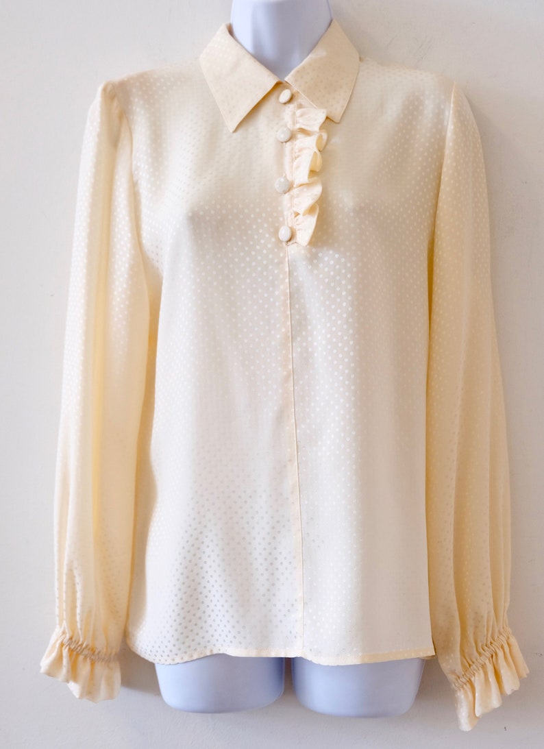 May include: A cream-colored, long-sleeved blouse with a ruffled collar and a button-up front. The blouse has a subtle polka dot pattern.