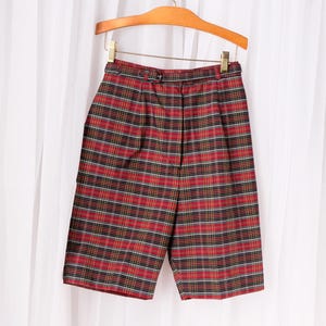 May include: Red, black, and gold plaid shorts hanging on a wooden hanger. The shorts have a high waist and a belt. The fabric is a woven material. The shorts are knee-length.