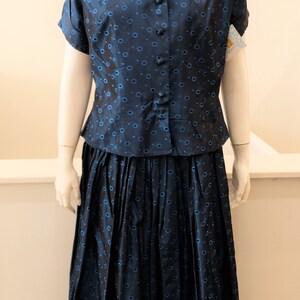 K&ouml;nnte beinhalten: Ein marineblaues zweiteiliges Vintage-Outfit. Das Set besteht aus einem kurz&auml;rmeligen Button-up-Hemd und einem plissierten Rock, beide mit einem sich wiederholenden blauen Blumenmuster. Der Stoff hat einen leichten Glanz.