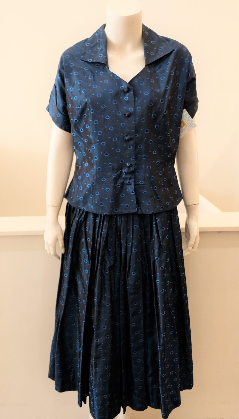 K&ouml;nnte beinhalten: Ein marineblaues zweiteiliges Vintage-Outfit. Das Set besteht aus einem kurz&auml;rmeligen Button-up-Hemd und einem plissierten Rock, beide mit einem sich wiederholenden blauen Blumenmuster. Der Stoff hat einen leichten Glanz.