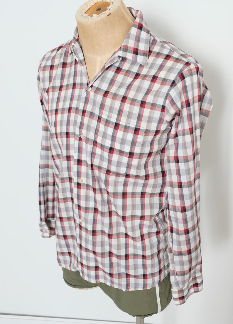 May include: A long-sleeved, button-down shirt with a classic plaid pattern in red, black, and gray on a white background. The shirt has a single chest pocket and is displayed on a mannequin.