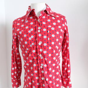 May include: A red shirt with a white polka dot pattern and small blue accents. The long-sleeved shirt has a collared neckline, two chest pockets, and white buttons. The shirt is displayed on a mannequin.