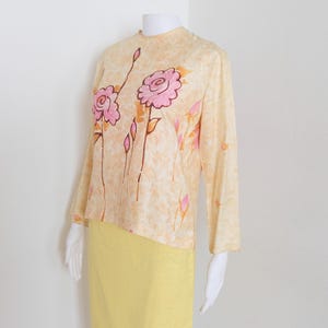 May include: A long-sleeved top in a pale peach hue with a mottled texture, featuring a hand-painted floral design of pink roses and orange buds on the front. The top is paired with a solid, bright yellow skirt, both displayed on a mannequin.