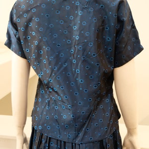 K&ouml;nnte beinhalten: Ein marineblaues zweiteiliges Outfit mit einem kurz&auml;rmeligen Oberteil mit Kragen und einem plissierten Rock. Der Stoff ist mit kleinen, hellblauen Blumenmustern versehen. Das Outfit wird auf einer Schaufensterpuppe pr&auml;sentiert.