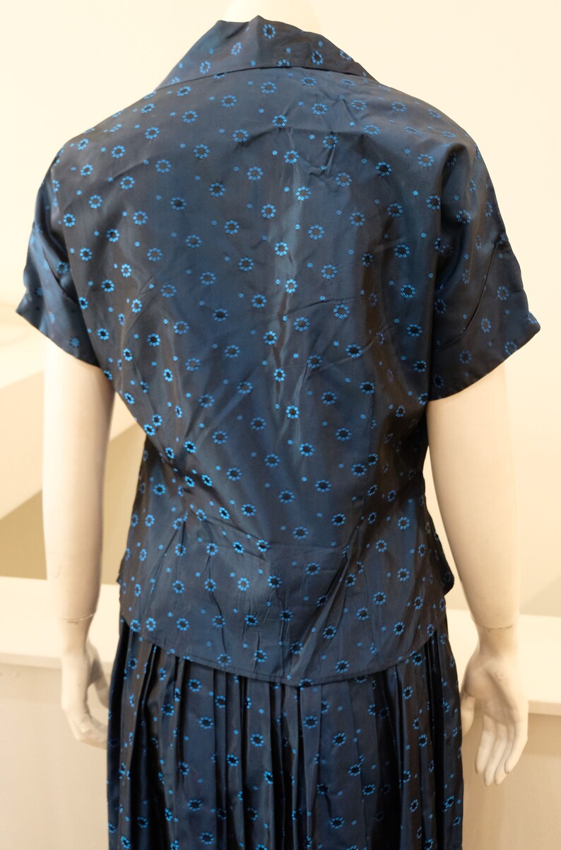 K&ouml;nnte beinhalten: Ein marineblaues zweiteiliges Outfit mit einem kurz&auml;rmeligen Oberteil mit Kragen und einem plissierten Rock. Der Stoff ist mit kleinen, hellblauen Blumenmustern versehen. Das Outfit wird auf einer Schaufensterpuppe pr&auml;sentiert.