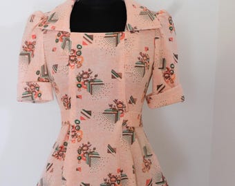 May include: A peach-colored, vintage-style top with a square neckline and short sleeves. The fabric features a repeating pattern of floral and geometric shapes in shades of brown, green, and orange. The top has a peplum-style waist and is displayed on a black mannequin.