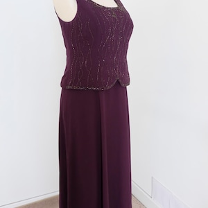May include: A sleeveless, floor-length, purple dress with a beaded bodice. The dress has a scoop neckline and a fitted bodice. The skirt is flowy and has a slight A-line shape.
