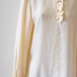 May include: A cream-colored, long-sleeved blouse with a white polka dot pattern. The blouse has a ruffled collar and cuffs, and features a row of white buttons down the front.