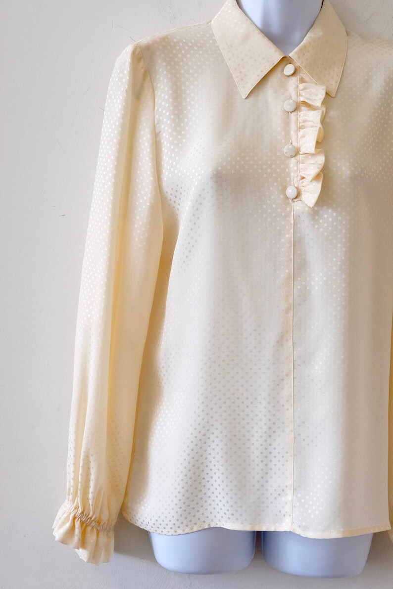 May include: A cream-colored, long-sleeved blouse with a white polka dot pattern. The blouse has a ruffled collar and cuffs, and features a row of white buttons down the front.