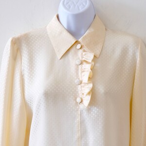 May include: A cream-colored, long-sleeved blouse with a white polka dot pattern and a ruffled trim around the collar.