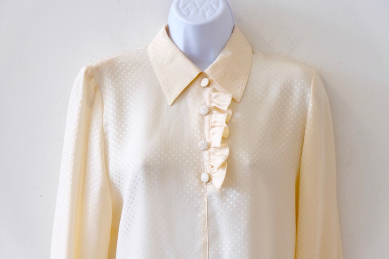 May include: A cream-colored, long-sleeved blouse with a white polka dot pattern and a ruffled trim around the collar.