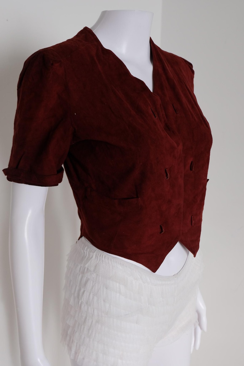 K&ouml;nnte beinhalten: Burgunderfarbene Wildlederjacke mit kurzen &Auml;rmeln und V-Ausschnitt. Die Jacke hat ein doppelreihiges Design mit mehreren kleinen Kn&ouml;pfen und zwei Vordertaschen. Die &Auml;rmel sind umgeschlagen und die Jacke wird auf einer wei&szlig;en Schaufensterpuppe pr&auml;sentiert.