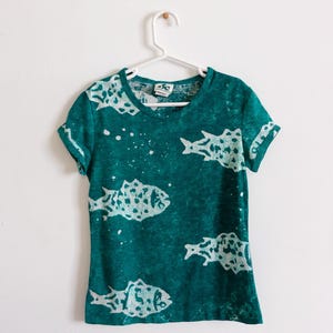 Op de afbeelding: Tealkleurig T-shirt met korte mouwen en een wit vispatroon. Het shirt hangt aan een witte hanger tegen een witte muur. Het visontwerp wordt herhaald over het shirt.