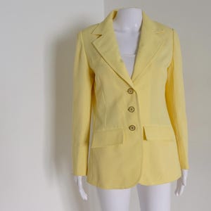 May include: A pale yellow blazer with a notched lapel, three gold-colored buttons, and two front pockets. The blazer is displayed on a white mannequin. The garment is a classic style.