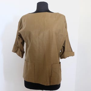 May include: A tan-colored top with a boat neck and three-quarter sleeves. The sleeves are cuffed. Two large pockets are visible on the front. The top is displayed on a black mannequin.