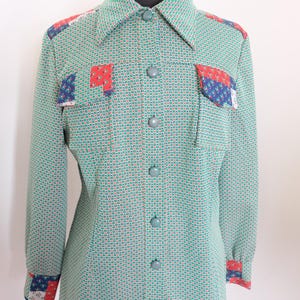 May include: A vintage green button-down shirt with a small square pattern. The shirt features patch pockets and cuffs with red, white, and blue patterned accents. The shirt is displayed on a mannequin.