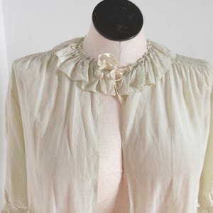May include: Pale green vintage robe with a ruffled collar and delicate bow detail. The robe has long sleeves and a flowing, open front design. The fabric appears lightweight and slightly sheer, suggesting a soft, comfortable feel. A classic piece of vintage sleepwear.