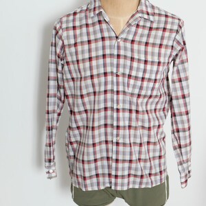 May include: A long-sleeved, button-down shirt with a plaid pattern in red, black, and white. The shirt is displayed on a mannequin and has a classic collar and a single chest pocket. The shirt is paired with olive green shorts.