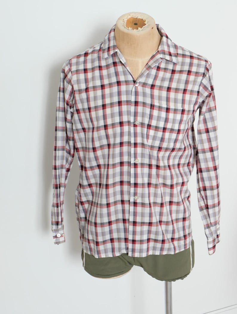 May include: A long-sleeved, button-down shirt with a plaid pattern in red, black, and white. The shirt is displayed on a mannequin and has a classic collar and a single chest pocket. The shirt is paired with olive green shorts.