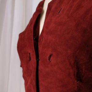 K&ouml;nnte beinhalten: Nahaufnahme einer bordeauxfarbenen Wildlederjacke. Die Jacke hat einen runden Ausschnitt, Knopfverschl&uuml;sse und kleine Taschen. Der Stoff hat ein weiches, strukturiertes Aussehen. Die Jacke wird auf einer Schaufensterpuppe pr&auml;sentiert.
