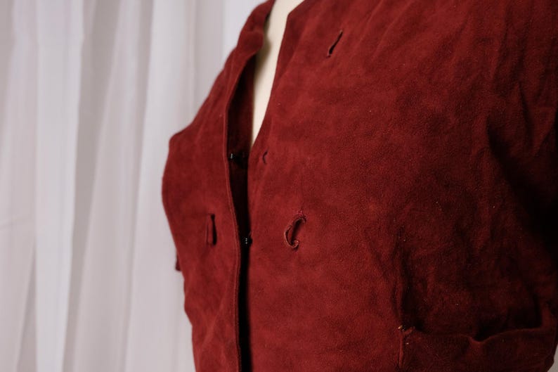 K&ouml;nnte beinhalten: Nahaufnahme einer bordeauxfarbenen Wildlederjacke. Die Jacke hat einen runden Ausschnitt, Knopfverschl&uuml;sse und kleine Taschen. Der Stoff hat ein weiches, strukturiertes Aussehen. Die Jacke wird auf einer Schaufensterpuppe pr&auml;sentiert.
