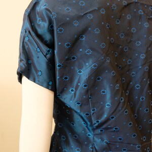 K&ouml;nnte beinhalten: Eine marineblaue Bluse mit kurzen &Auml;rmeln und einem floralen Muster in einem helleren Blau. Der Stoff hat einen dezenten Glanz. Die Bluse ist Teil eines Sets, mit einem passenden Rock unten sichtbar.