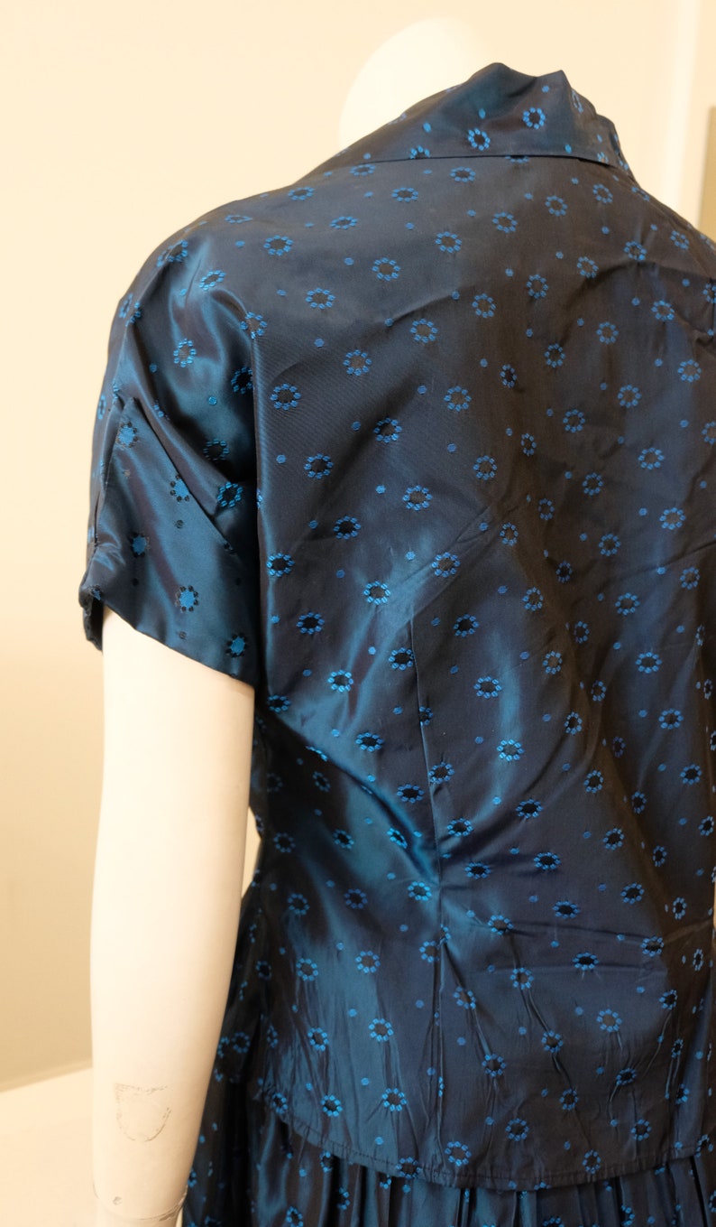 K&ouml;nnte beinhalten: Eine marineblaue Bluse mit kurzen &Auml;rmeln und einem floralen Muster in einem helleren Blau. Der Stoff hat einen dezenten Glanz. Die Bluse ist Teil eines Sets, mit einem passenden Rock unten sichtbar.
