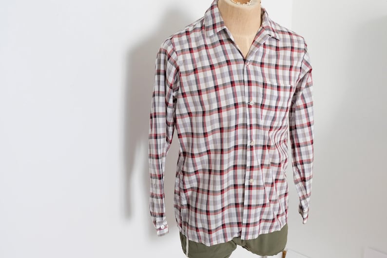 May include: A long-sleeved, button-down shirt in a classic plaid pattern. The shirt features a gray, red, and white checkered design. The shirt is displayed on a mannequin, showcasing its fit and style. The garment has a collared neckline and buttoned cuffs.