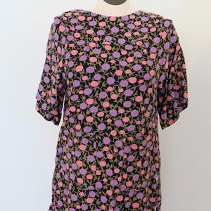 May include: A black floral print tunic-style top with short sleeves. The pattern features pink and purple flowers with green stems and leaves. The top is displayed on a mannequin against a white background.
