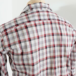 May include: A plaid button-down shirt in a gray, white, and red checkered pattern. The long-sleeved shirt is displayed on a wooden hanger against a white background. The back of the shirt is visible.