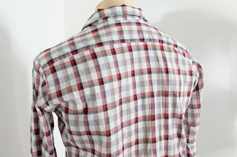 May include: A plaid button-down shirt in a gray, white, and red checkered pattern. The long-sleeved shirt is displayed on a wooden hanger against a white background. The back of the shirt is visible.