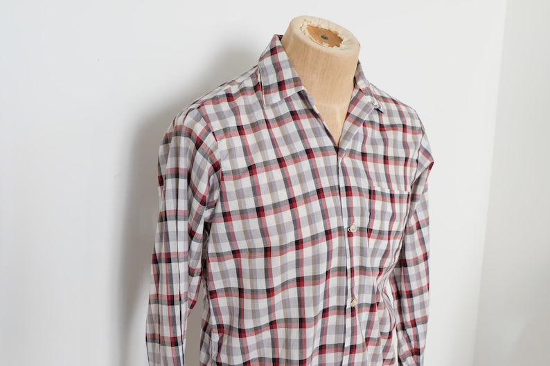 May include: A long-sleeved plaid shirt in a gray, red, and black pattern. The shirt is buttoned and has a collar. The shirt is displayed on a mannequin.