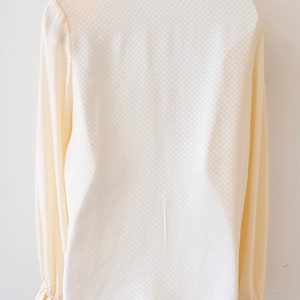 May include: A cream-colored long-sleeved blouse with a white polka dot pattern. The blouse has a ruffled collar and cuffs.