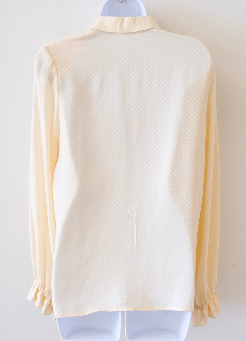 May include: A cream-colored long-sleeved blouse with a white polka dot pattern. The blouse has a ruffled collar and cuffs.