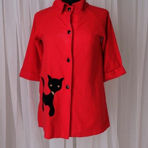 May include: A bright red jacket with a black cat applique on the front. The jacket has a collared neckline, three-quarter sleeves, and black buttons. The black cat has gold-colored eyes.