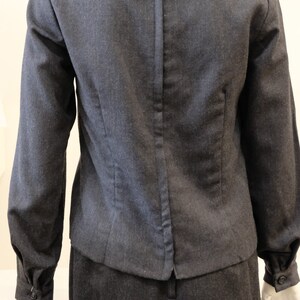 May include: A dark gray skirt suit with a long-sleeved jacket featuring a wide collar. The jacket has a zipper closure in the back.