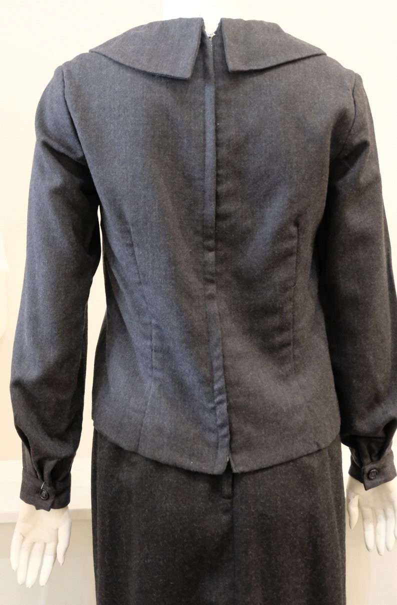 May include: A dark gray skirt suit with a long-sleeved jacket featuring a wide collar. The jacket has a zipper closure in the back.