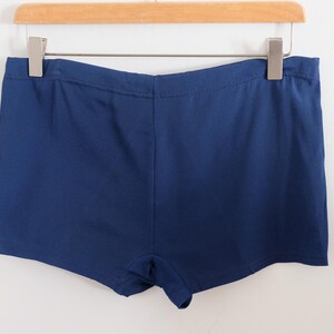 May include: A pair of navy blue shorts hangs from a wooden hanger against a white wall. The shorts are made of a solid, dark blue fabric. The shorts have a simple, classic design, suitable for casual wear.