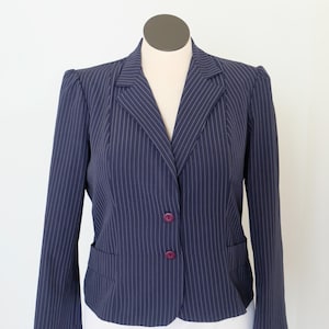May include: Navy blue pinstripe blazer with two purple buttons and two front pockets. The blazer is displayed on a mannequin and has a classic, tailored design. The white pinstripes add a subtle contrast to the dark blue fabric.