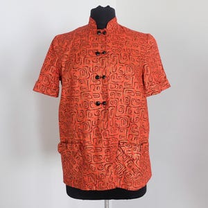 May include: Orange short-sleeved shirt with a mandarin collar and black buttons. The shirt features a repeating black abstract pattern. The shirt is displayed on a black mannequin.